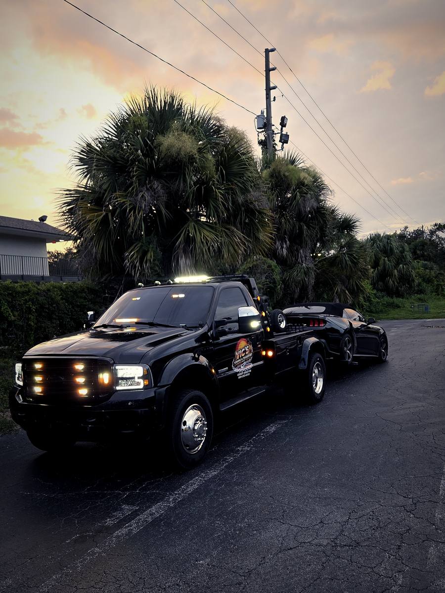 Vic's black Ford wrecker towing a Mustang convertible at sunset, palm trees behind.