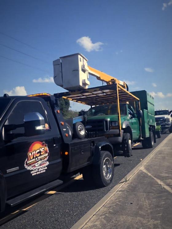 A bucket truck being towed by Vic's wrecker on a highway shoulder.
