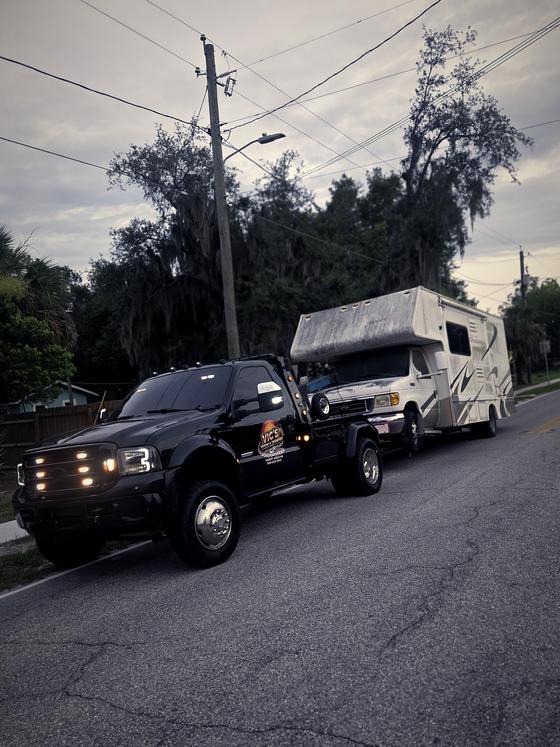 A Class C motorhome under tow behind Vic's wrecker on a residential street.